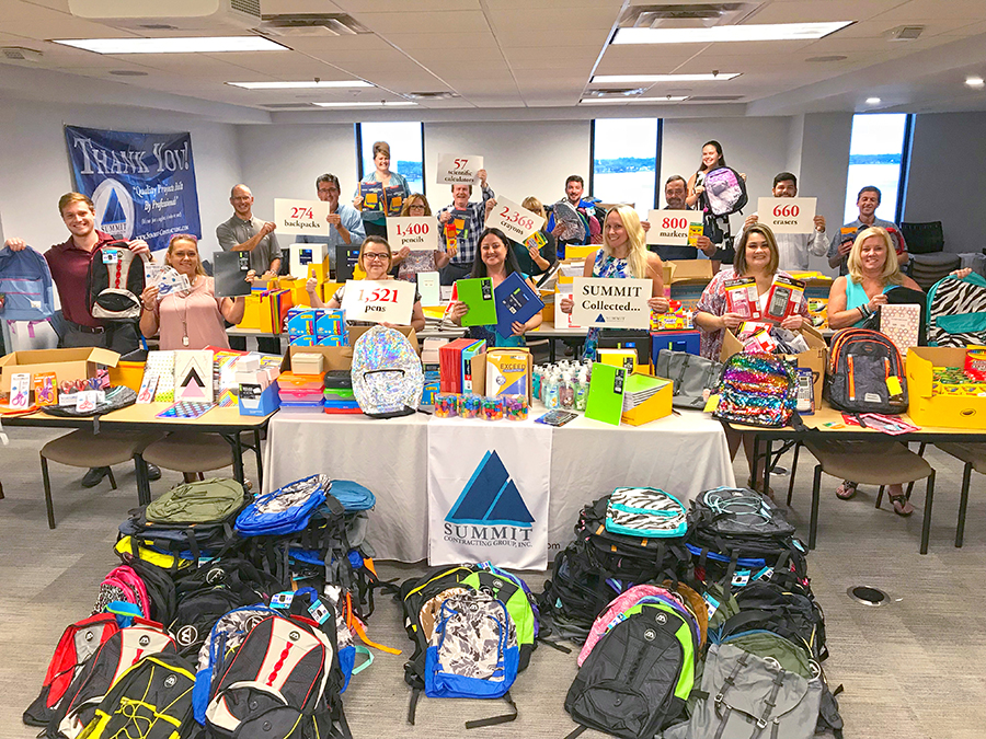 Backpacks and school supplies collected by Summit employees holding signs with data - 274 backpacks, 1,400 pencils, 2,368 crayons, 1,521 pens, 800 markers, 600 erasers, 57 scientific calculators collected by Summit Contracting Group